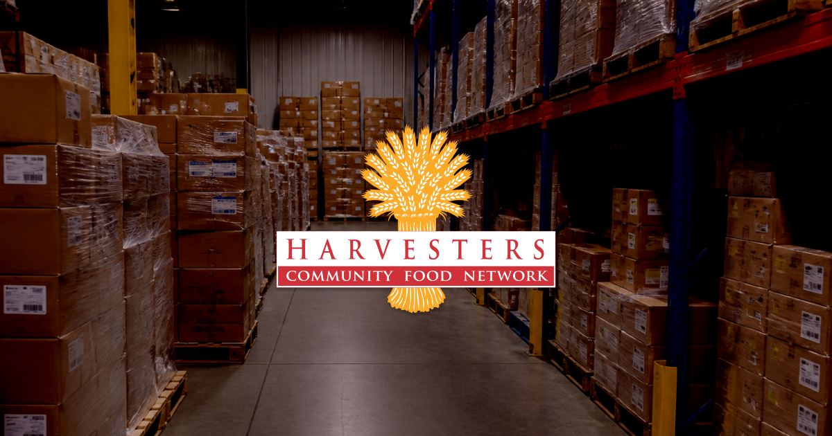 A woman stands in the foreground at a Harvesters food distribution; behind her, a volunteer wheels a cart of squash down a truck ramp and people wait in line in the sun.