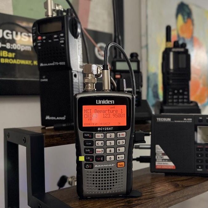 A Uniden BC125AT handheld scanner mid-tune, reading 'MCI Departure 1, 123.9500,' on a wood shelf.