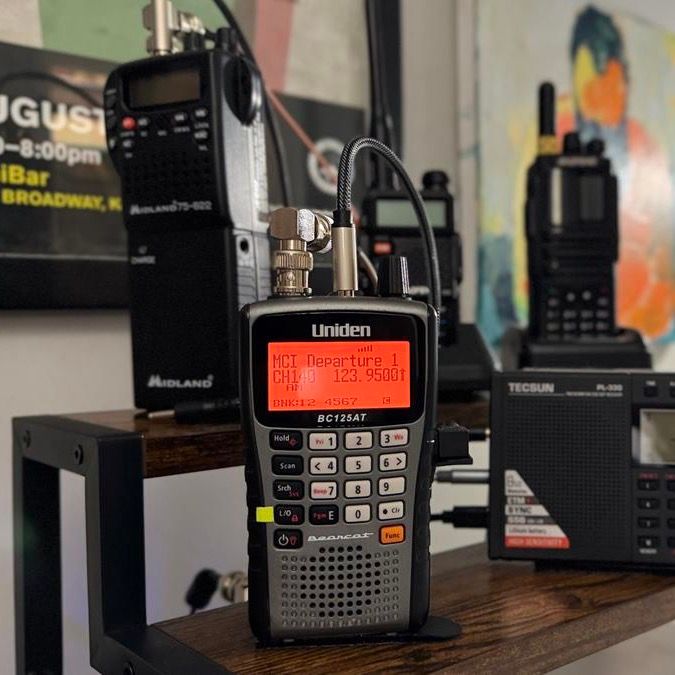 A Uniden BC125AT handheld scanner mid-tune, reading 'MCI Departure 1, 123.9500,' on a wood shelf.