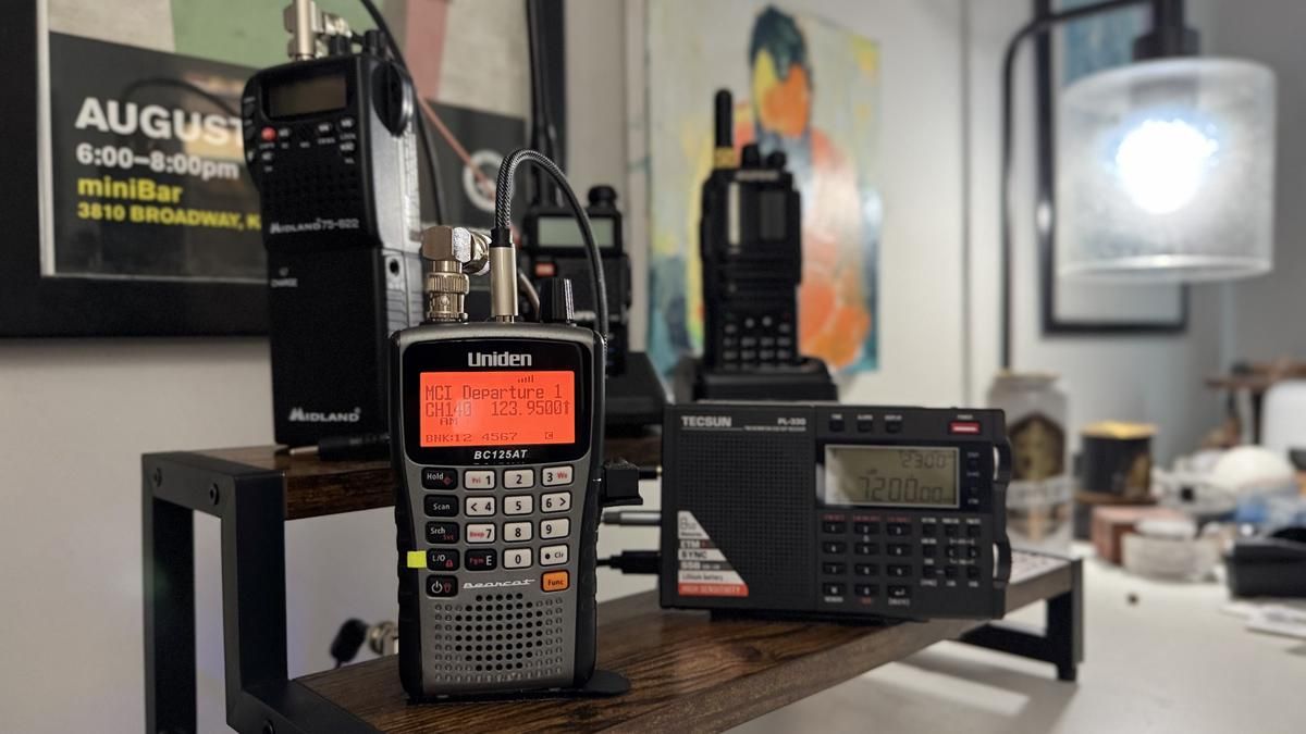 A handheld radio bench: a Uniden BC125AT scanner lit up in front, with a Midland weather radio and a Tecsun PL-330 shortwave receiver behind it, on a wood shelf.