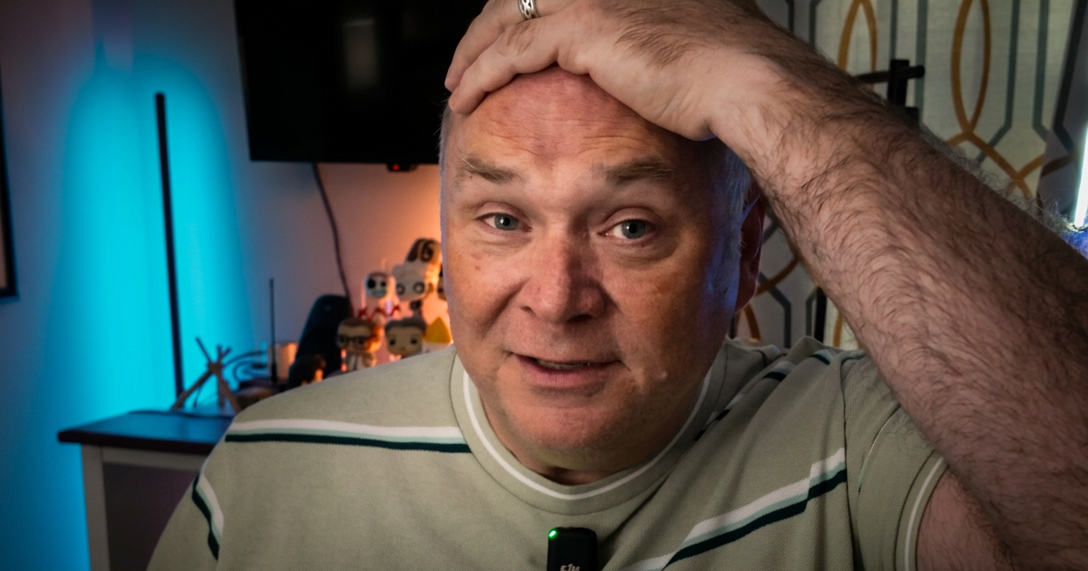 Close-up video still of me at my desk, one hand pushed up over my forehead, a lavalier mic clipped to my striped polo, the room behind me lit teal.