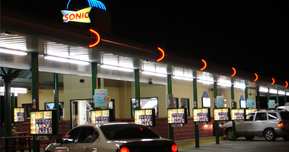 A Sonic Drive-In at night, two cars nosed into glowing menu-board stalls beneath the lit neon sign.